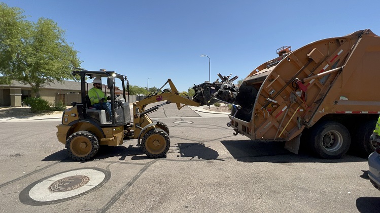 A bulk trash equipment operator uses a tractor to lift large items into a rear-loader garbage truck.