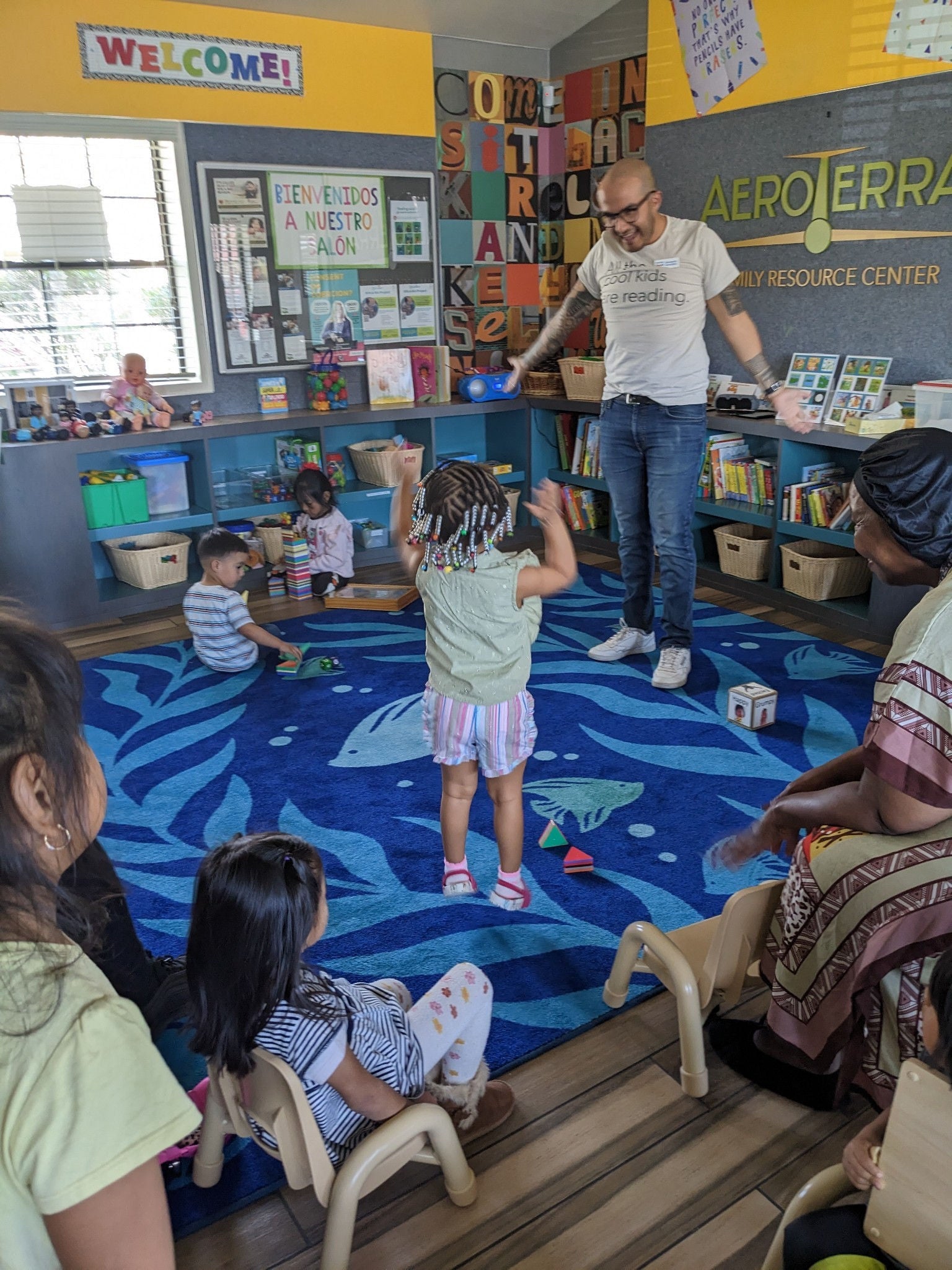 In what appears to be a (very colorful) elementary school classroom, a young student with her back to the camera appears to be jumping up and down while her adult teacher looks on with a happy expression with his arms outstretched. Other students are seated around the area -- some on a blue rug and others in small chairs.