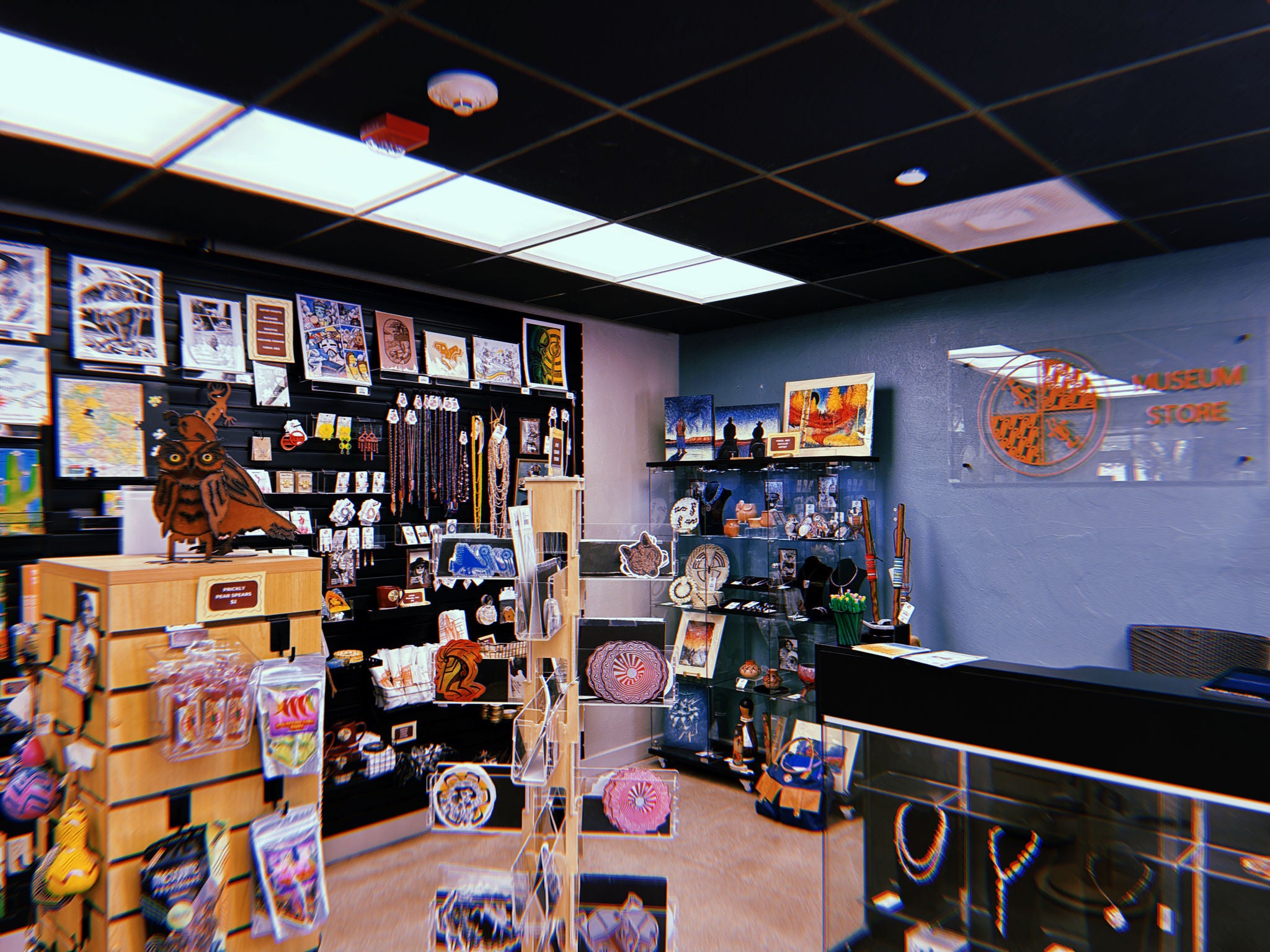 A view inside the S’edav Va’aki Museum Store showing a well‑lit retail space filled with Southwestern and Indigenous‑themed merchandise. The left side features wall displays with art prints, postcards, ornaments, and hanging beaded items. Freestanding racks hold stickers, small crafts, and packaged goods. In the center foreground is a wooden display with a carved owl figure perched on top. On the right side, a glass jewelry case contains necklaces, while shelves behind it display pottery, figurines, and artwork. A transparent wall sign reads “Museum Store,” with a circular orange logo. The space has dark ceiling tiles, bright fluorescent lighting, and neutral‑colored floors.