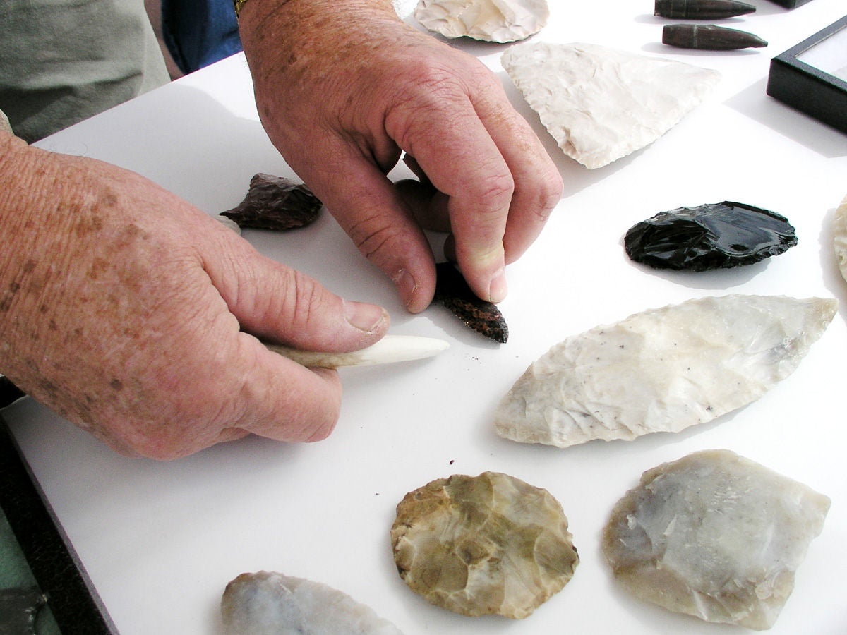 Photo of hands using antler billet to flint knap a projectile point with other sherds in the foreground. 