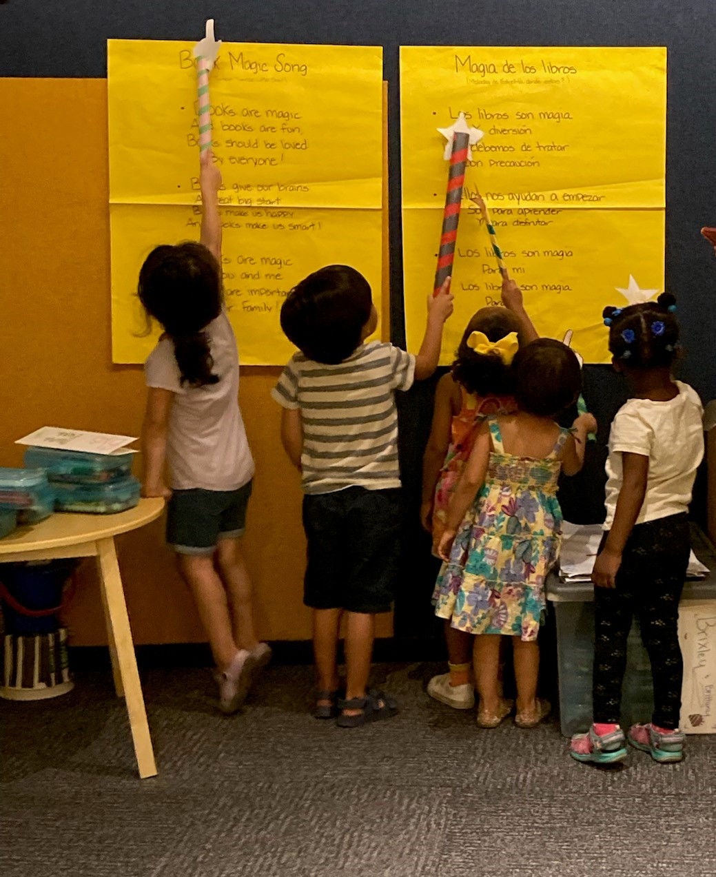 Five young students in preschool or elementary school stand together in front of a wall with two yellow posters on it. The posters have both Spanish and English song lyrics written on them. The children are using homemade paper wands to point to certain words. The setting is a classroom.