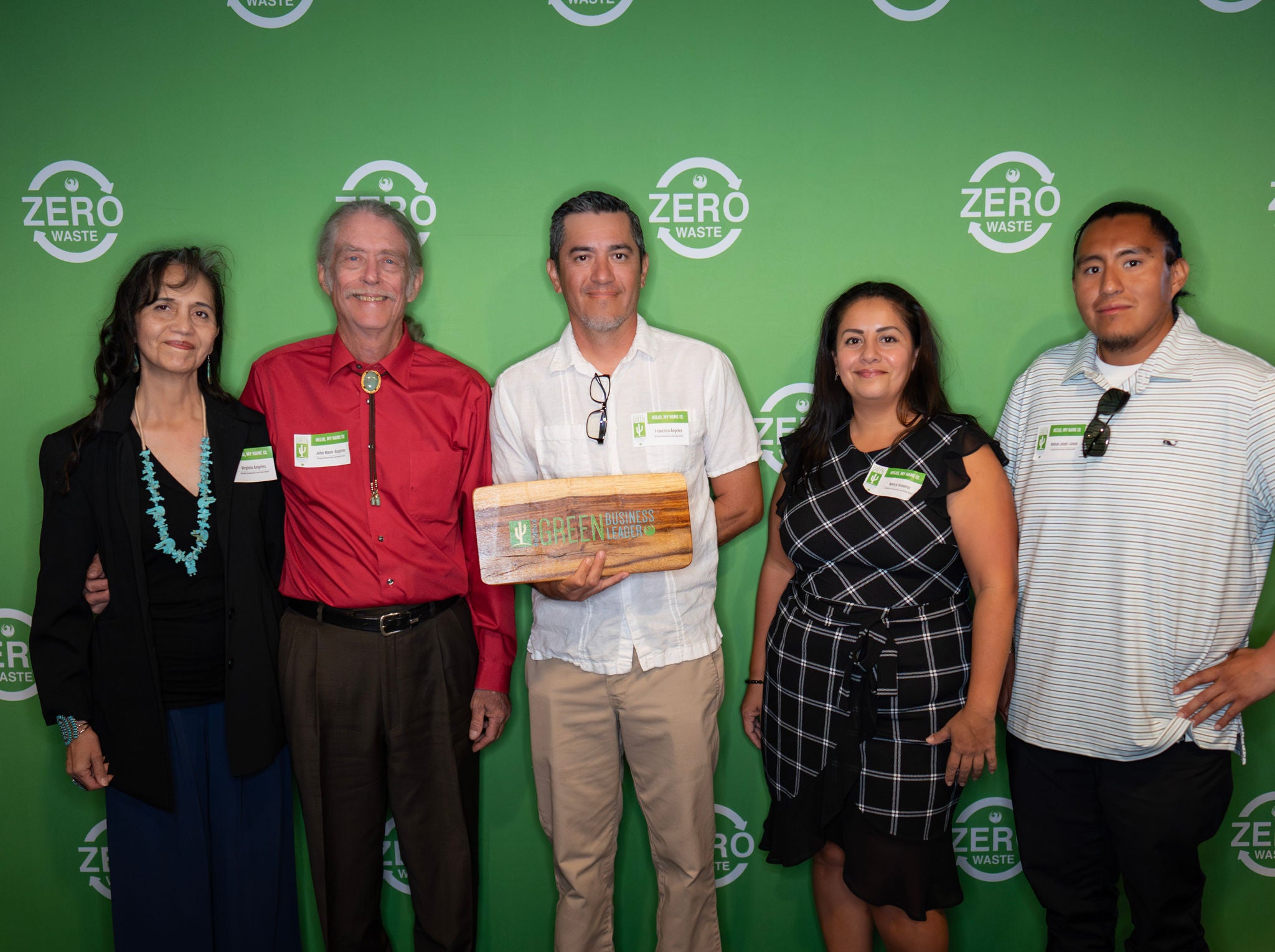 A group of new Green Business Leaders holds their recognition plaque in front of a green backdrop with the Zero Waste logo all over it
