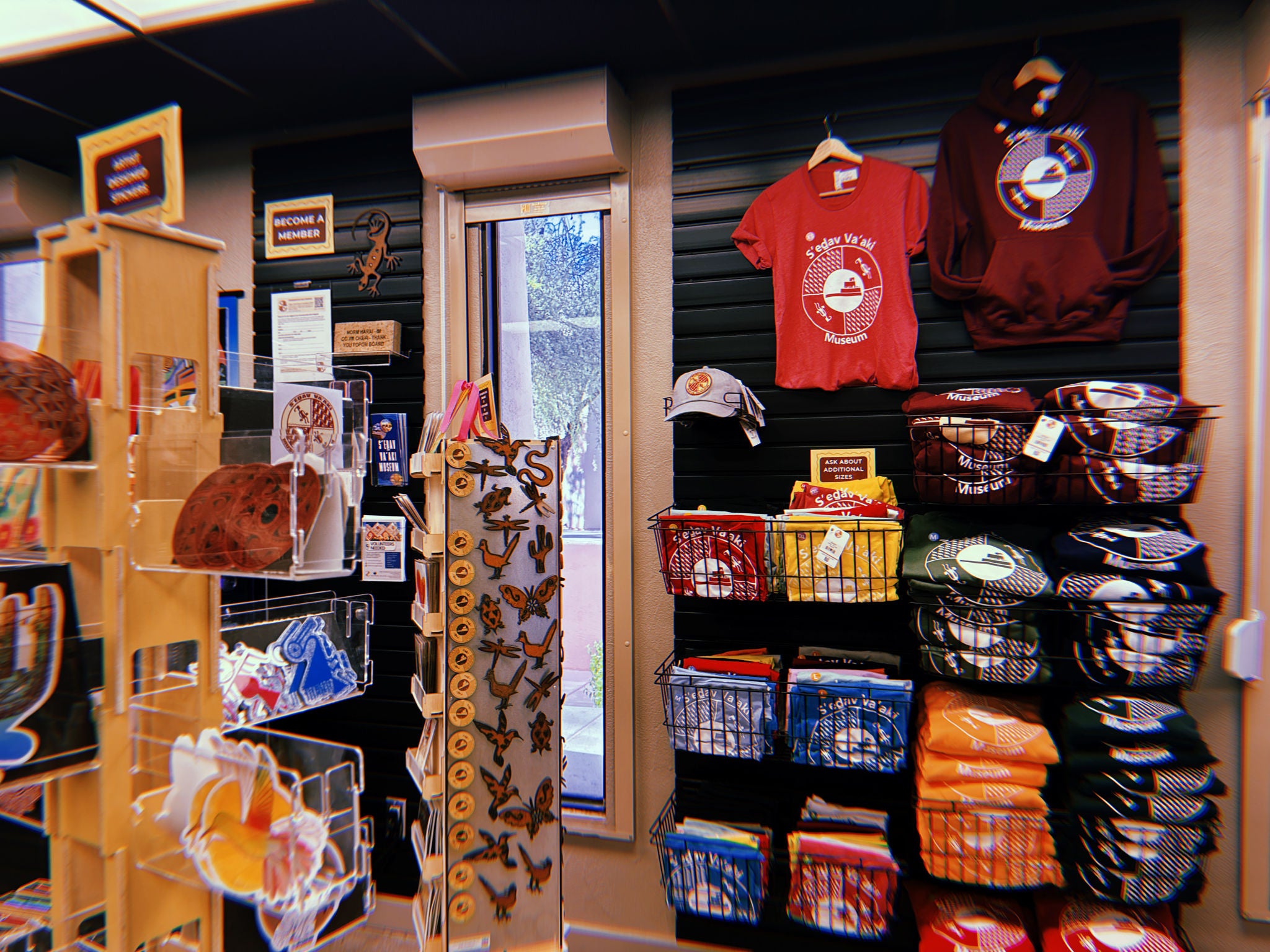A portion of the S’edav Va’aki Museum Store featuring apparel and small souvenirs. On the right wall, wire baskets hold folded T‑shirts in multiple colors, all printed with the museum’s circular logo. Above them, a red T‑shirt and a maroon hoodie with the same logo hang on display. A gray cap is mounted below. In the center, a rotating stand holds wooden magnets and ornaments shaped like desert animals and symbols. On the left, clear acrylic racks hold postcards and artwork. A tall narrow window shows outdoor trees and daylight. Overhead fluorescent lighting illuminates the area.