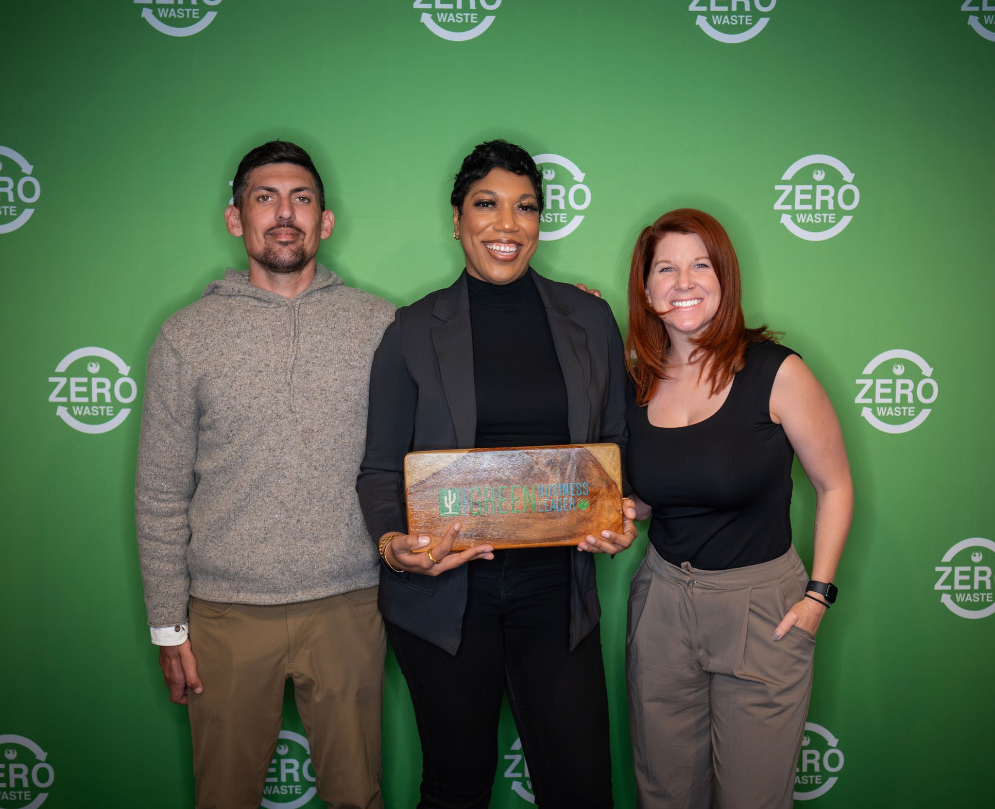 A new Green Business Leader holds his recognition plaque in front of a green backdrop with the Zero Waste logo all over it