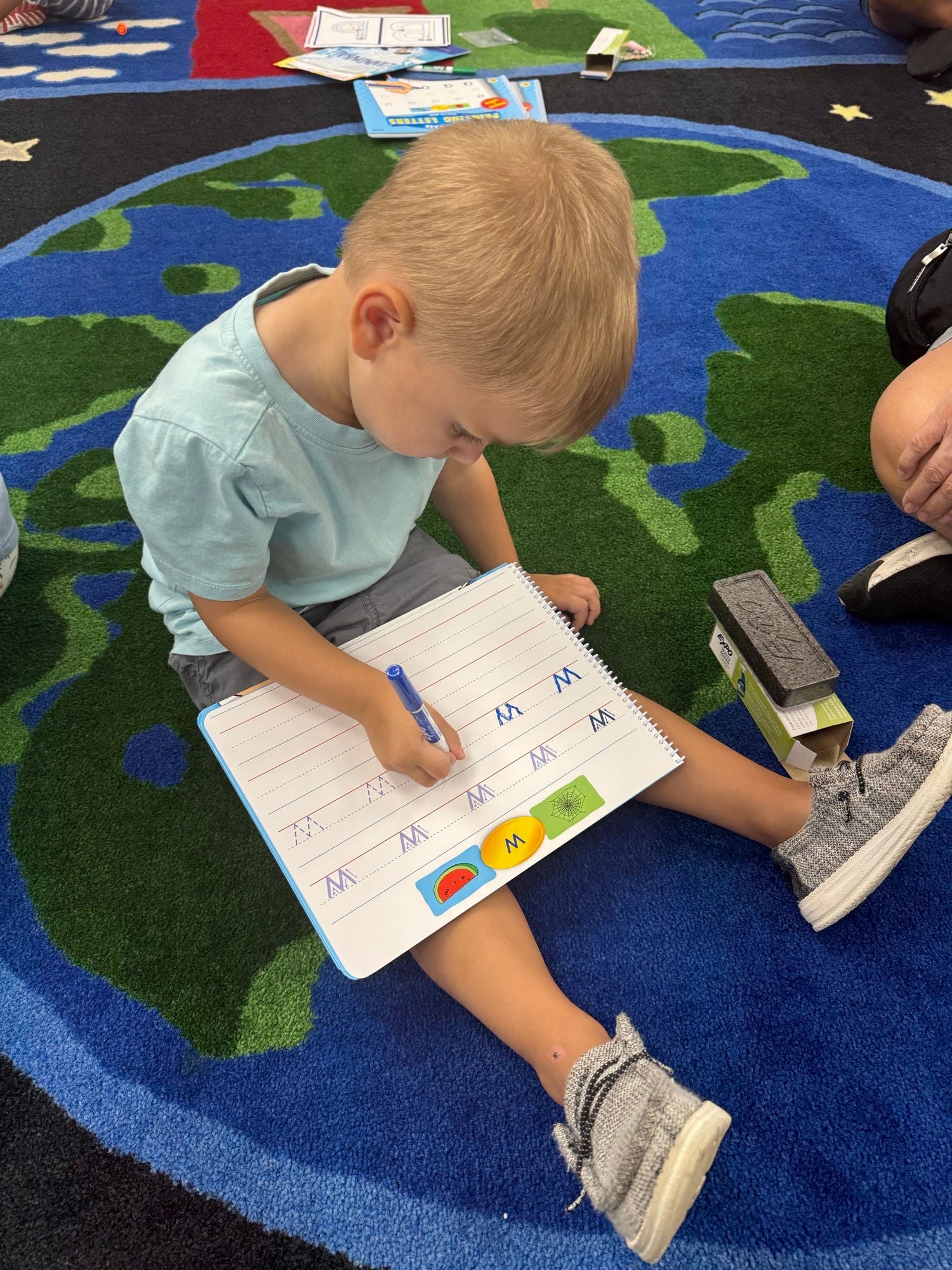 A toddler sits on the floor on a bright blue and green rug. The toddlers legs are in front of him, and in his lap is a white board on which he is practicing writing the letter 'W.'