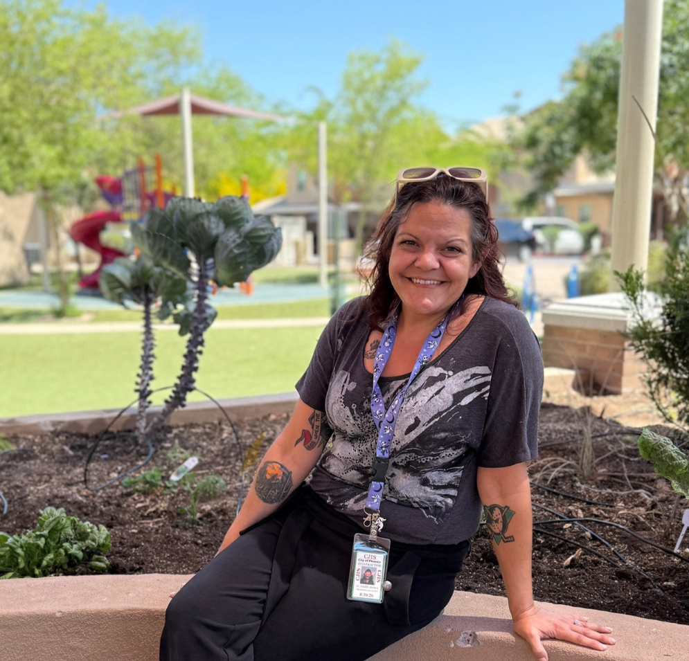 A woman smiles as she sits on the edge of a raised flower bed at a park. It is sunny with blue skies.