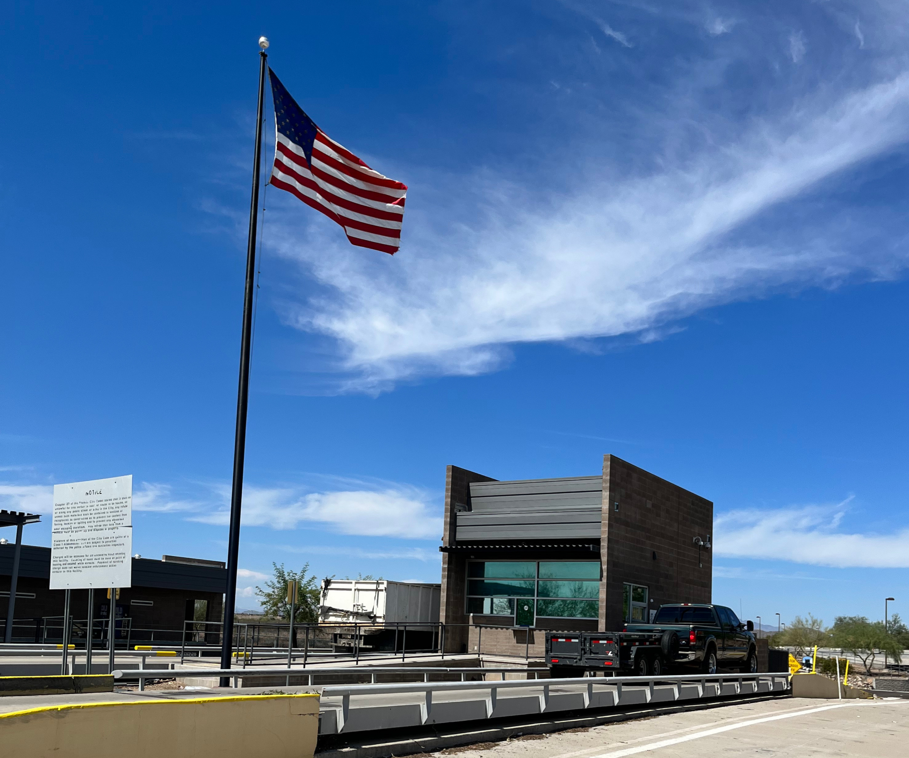 A black pickup truck next to the window of the 27th Avenue Scale House at the entrance of the 27th Avenue Transfer Station. A vividly blue sky with some thin clouds is seen during afternoon daylight. An American flag is waving at the top of a flag pole next to the scale house.