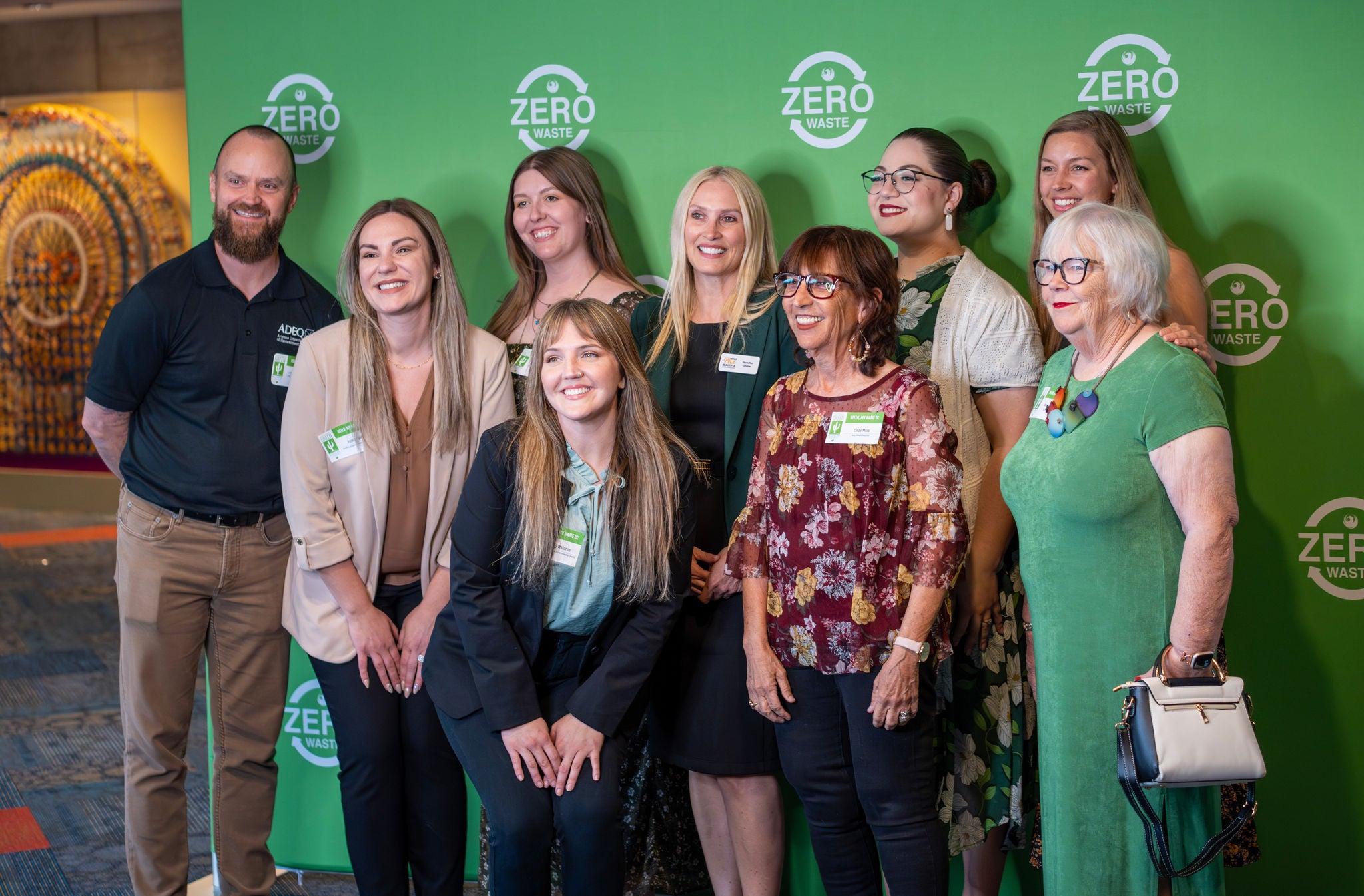 Guests representing a certified Green Business Leader stand for a photo opp in front of a green backdrop that is filled with the Zero Waste logo..