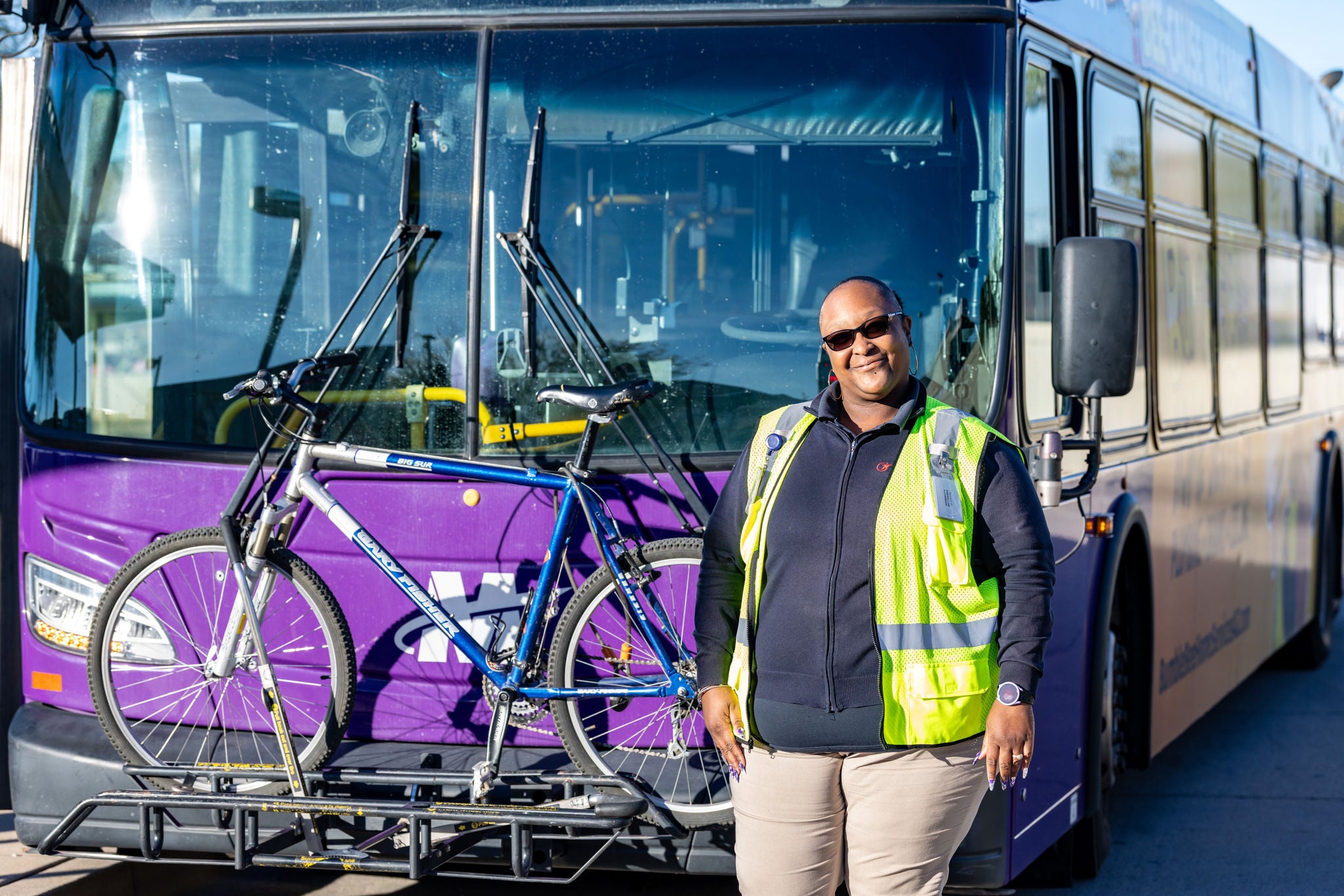 Female bus driver standing in front of a bus while posing and smiling. 