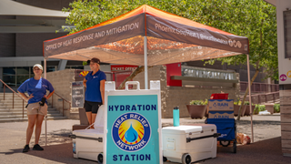 Two individuals standing under a tent with a hydration station sign in front of them 