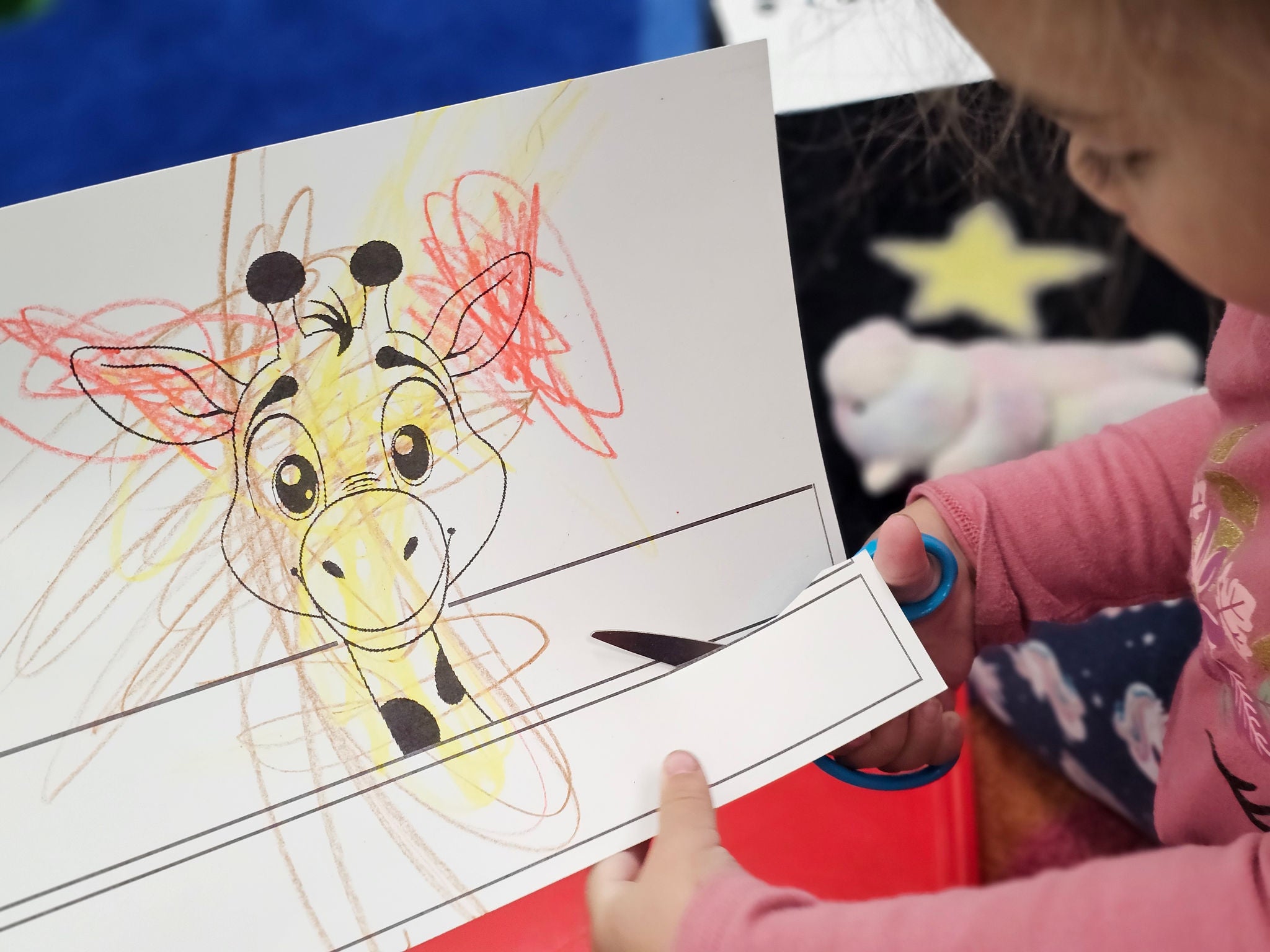 An preschool or early elementary school student uses scissors to cut a piece of paper which has a cartoon giraffe on it. The giraffe appears to have been colored by a child.