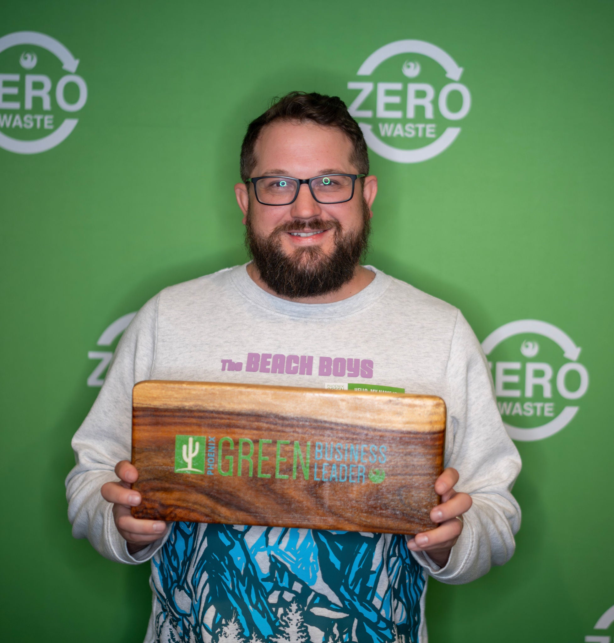 A new Green Business Leader holds his recognition plaque in front of a green backdrop with the Zero Waste logo all over it