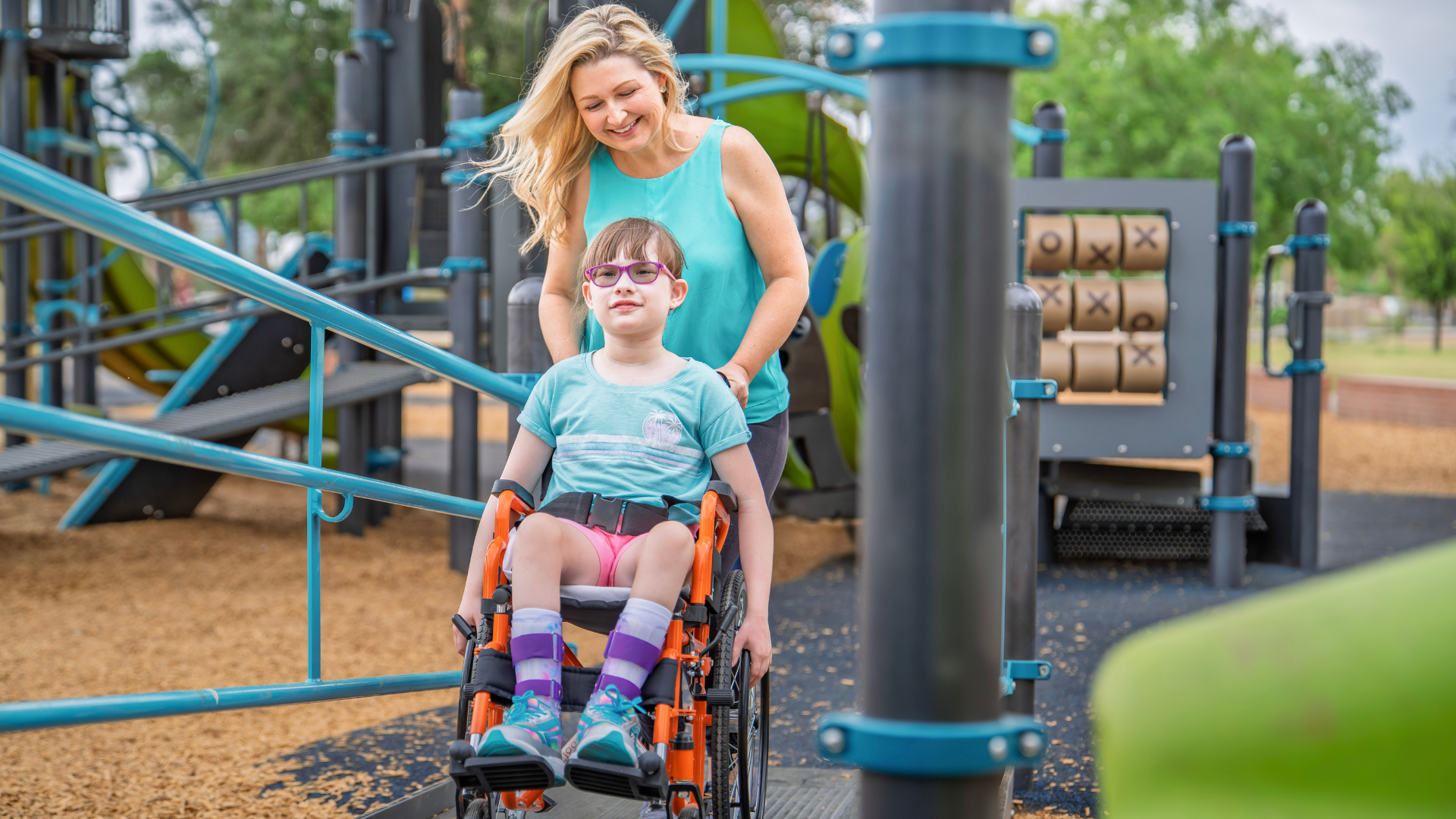 Mother pushing daughter in mobility device along a path at Encanto Park with playground in the background