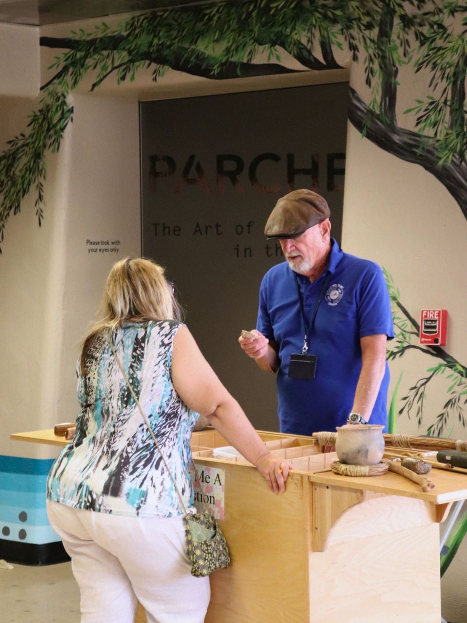 An Engagement Guide volunteer at the S’edav Va’aki Museum stands behind the Artifact Cart, speaking with a visitor who is standing on the opposite side of the wooden display. The cart holds several replica tools and artifacts. Behind them is a wall with a painted mural of tree branches and a partially visible title from a past traveling exhibit, “Parched: The Art of Water in the Southwest.”