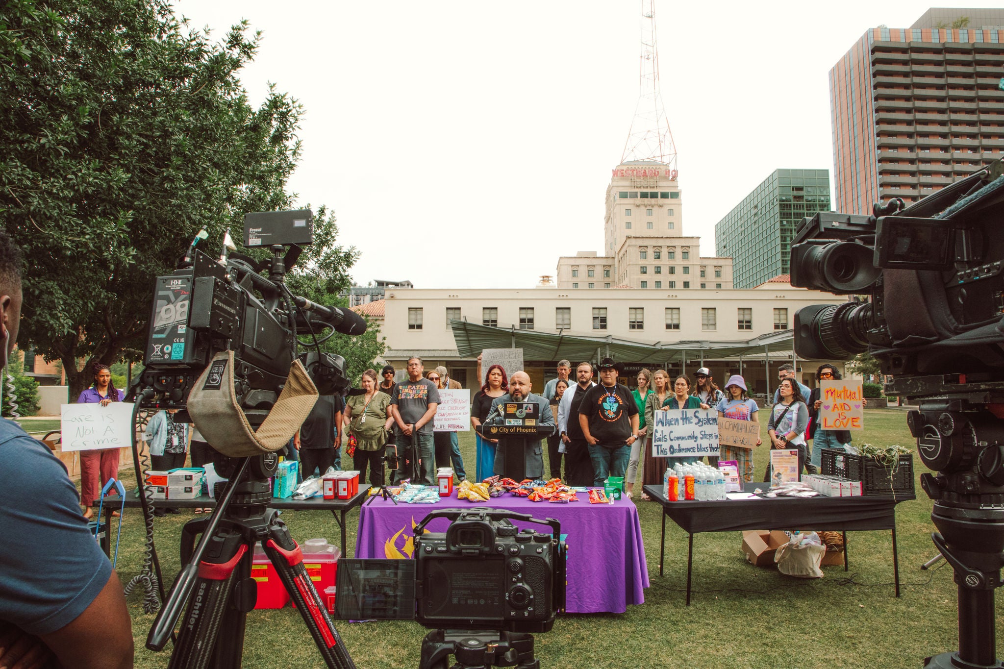 A man speaks at a podium marked 'City of Phoenix' during an outdoor press conference, surrounded by community members holding signs reading 'Care Is Not A Crime,' 'When the System Fails Community Steps In,' 'Decriminalize,' and 'Mutual Aid Is Love.' Tables in front of the group hold mutual aid supplies including food, water, and first aid items. News cameras are set up in the foreground, with downtown Phoenix buildings visible in the background.