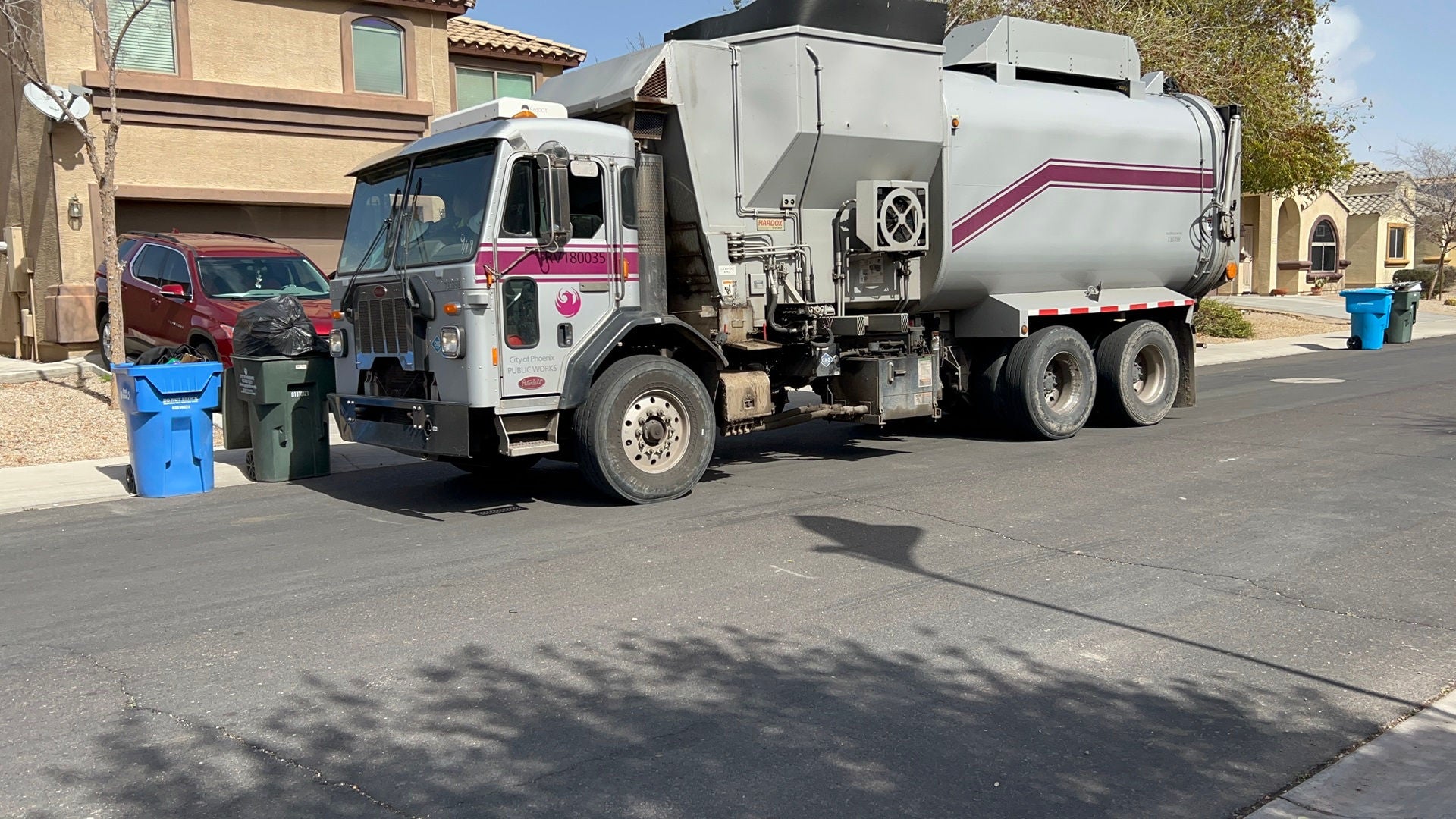A silver City of Phoenix garbage truck collects trash from curbside containers.