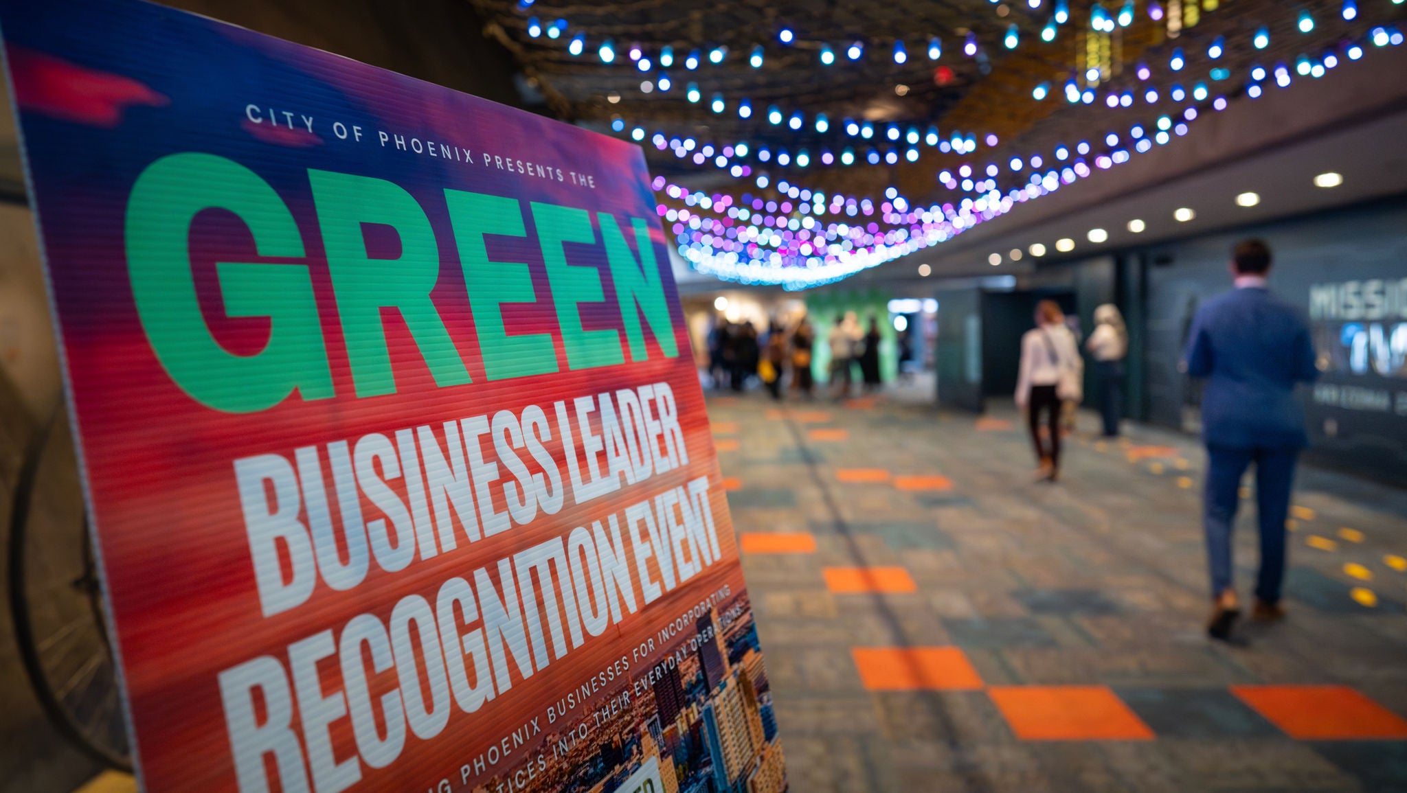 A colorful sign reads 'Green Business Leader Recognition Event' as people walk by.
