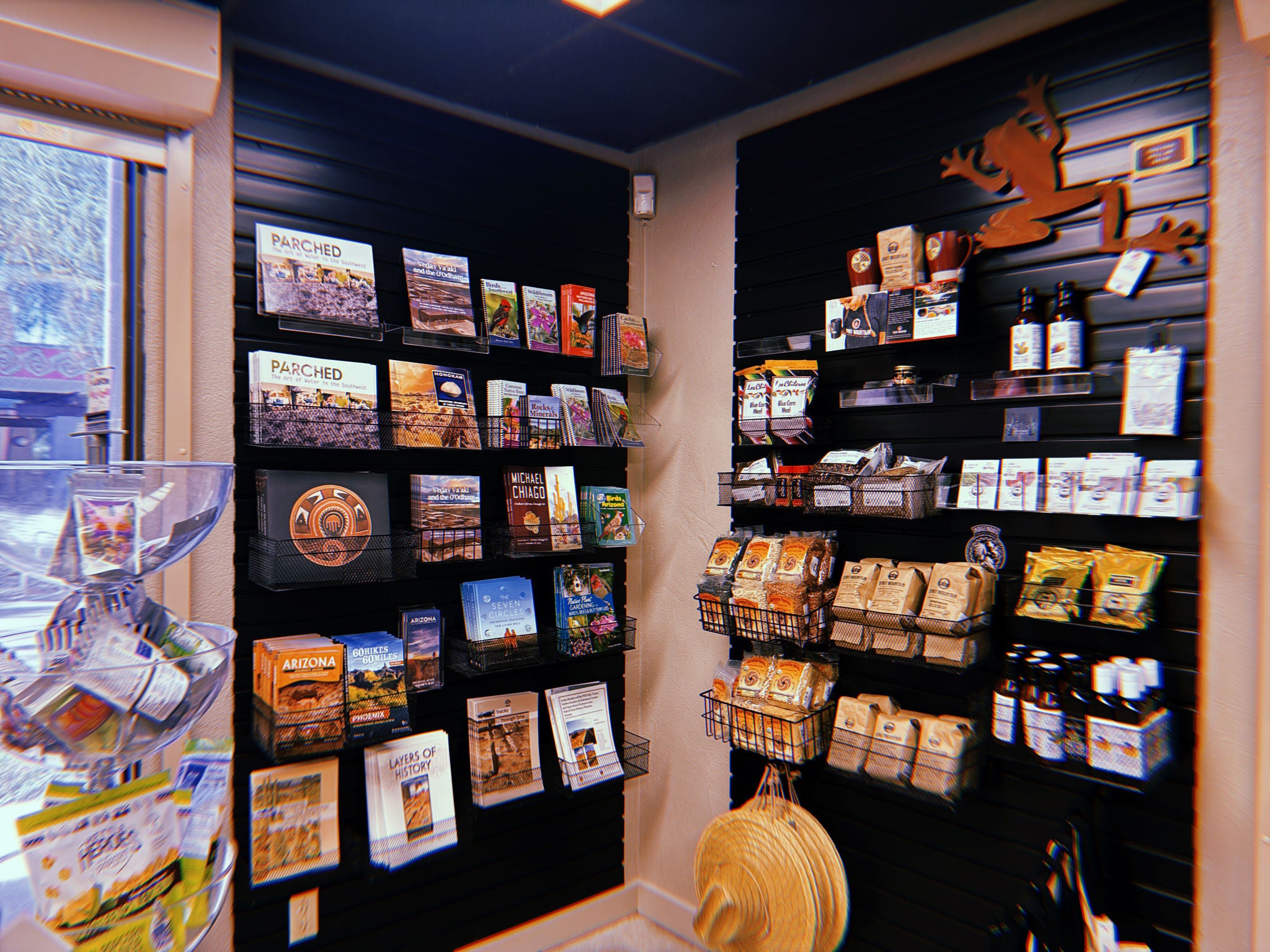 A section of the S’edav Va’aki Museum Store showing two black slat‑wall displays. The left wall holds books and guidebooks arranged on shelves and in wire baskets. The right wall displays packaged food items, coffee bags, bottled products, and small gift items. A metal lizard decoration is mounted near the top. On the far left, a rotating stand with clear bins holds small souvenirs. A window on the left lets in daylight, and the area is lit by overhead fluorescent lights.