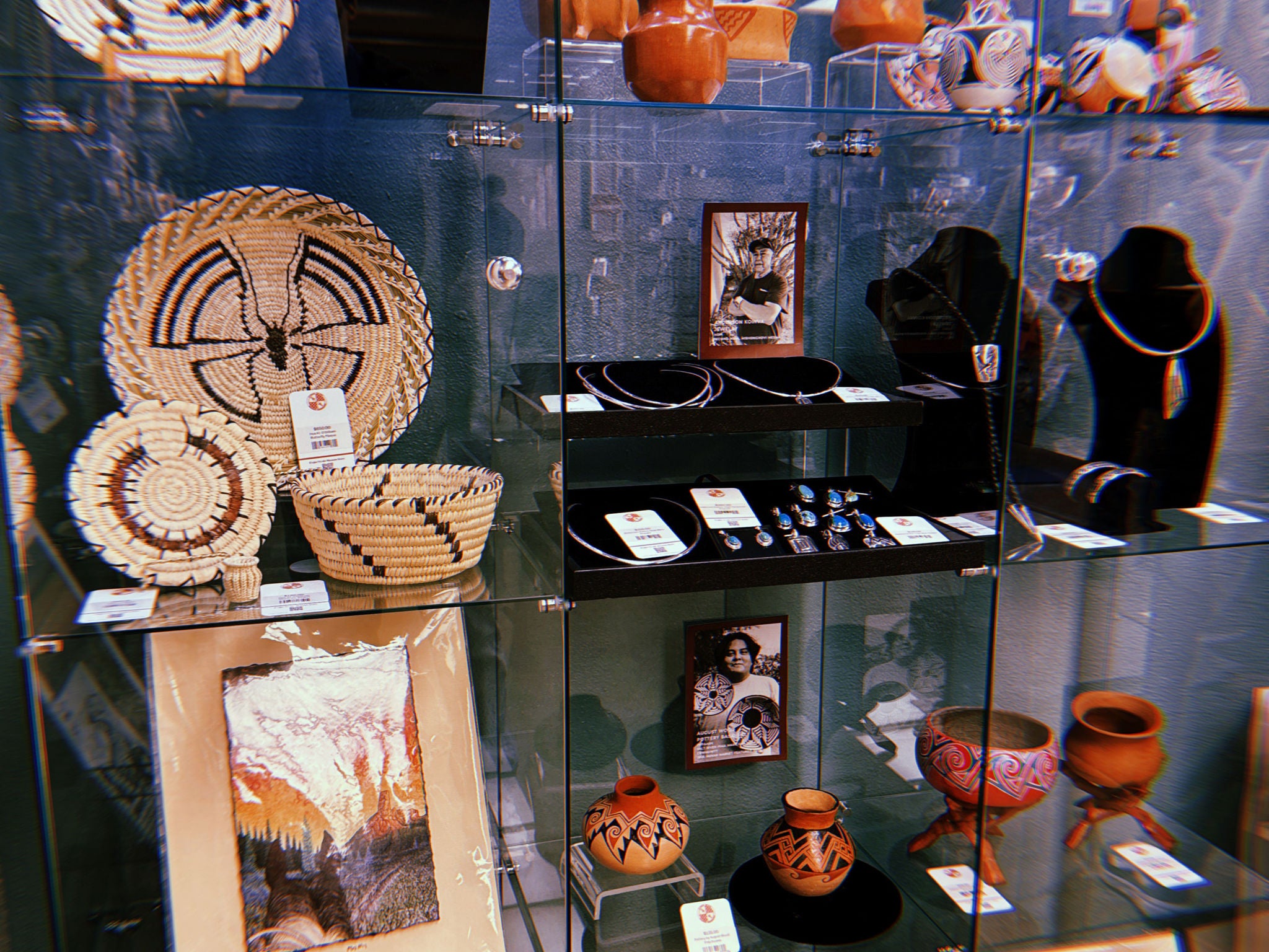 A glass display case in the S’edav Va’aki Museum Store containing woven baskets, jewelry, pottery, and framed artwork. On the left, several coiled baskets with geometric patterns sit on a glass shelf. In the center, black necklace stands display silver and beaded jewelry, with rings arranged on a lower shelf. The right side holds painted pottery bowls and vessels with geometric designs. Framed artwork is placed both inside the case and on the shelf below. The items are arranged neatly on multiple glass shelves.