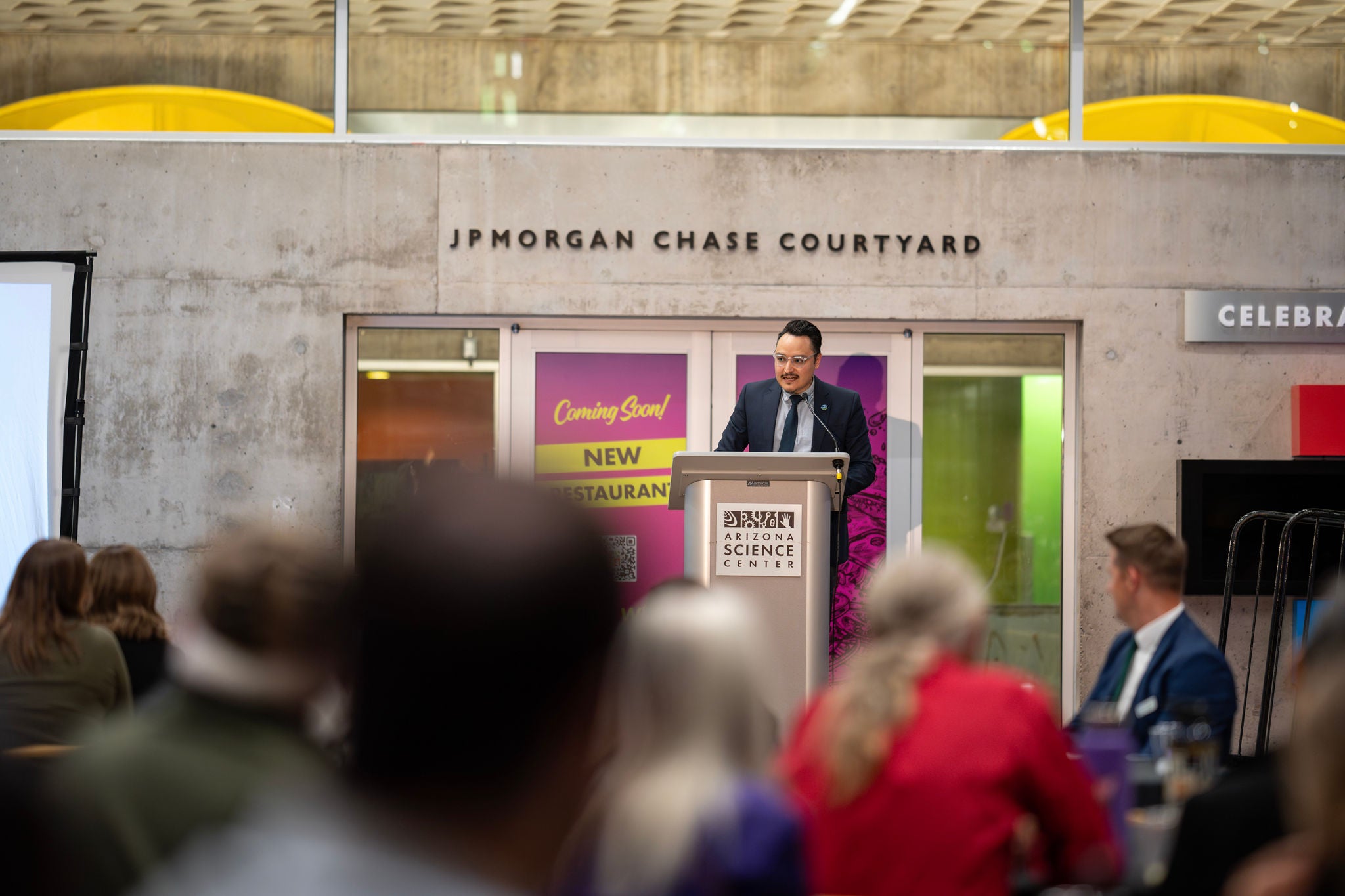 The crowd seated around tables at the Arizona Science Center (foreground) with a speaker at the podium in focus in the background.
