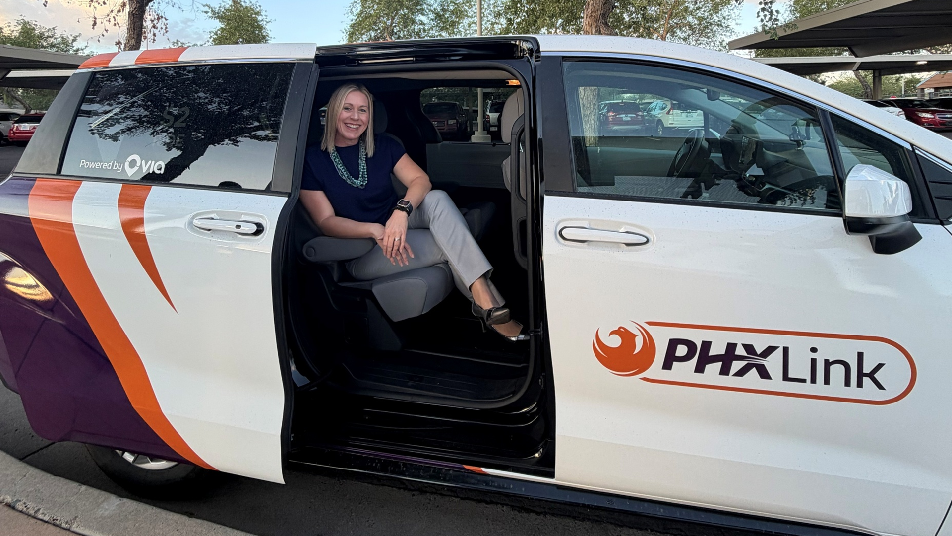 Image of a woman sitting in the back seat of a PHXLink vehicle. The vehicle is a white van with orange and purple designs. You can see the PHXLink logo on the front passenger seat door. 