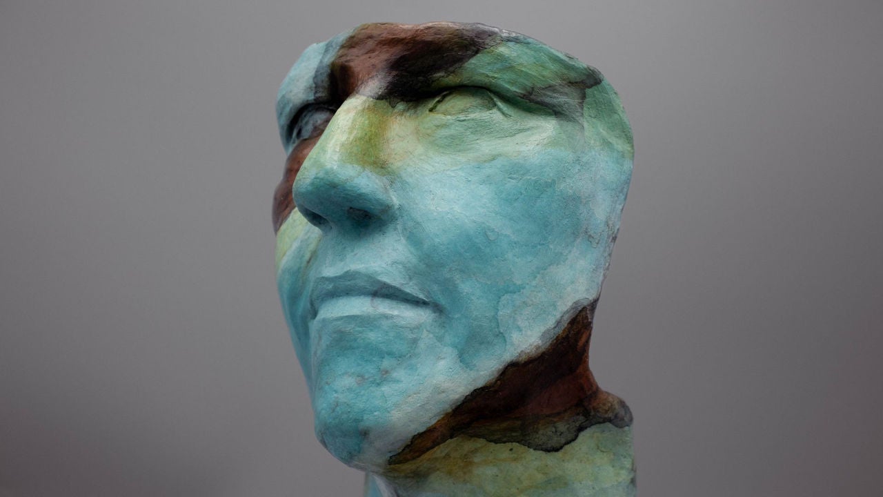 Photo of Holisso Pisachi' by Addison Karl. A bust casted in bronze, oxidized to show various colors including but not limited to teal, green, yellow, and brown. 