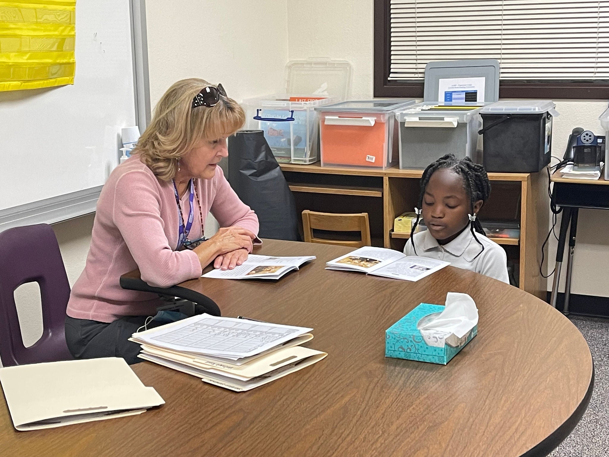 An adult and a young student sit together at a semi-circular table in an elementary school classroom. They each have a booklet open in front of them.