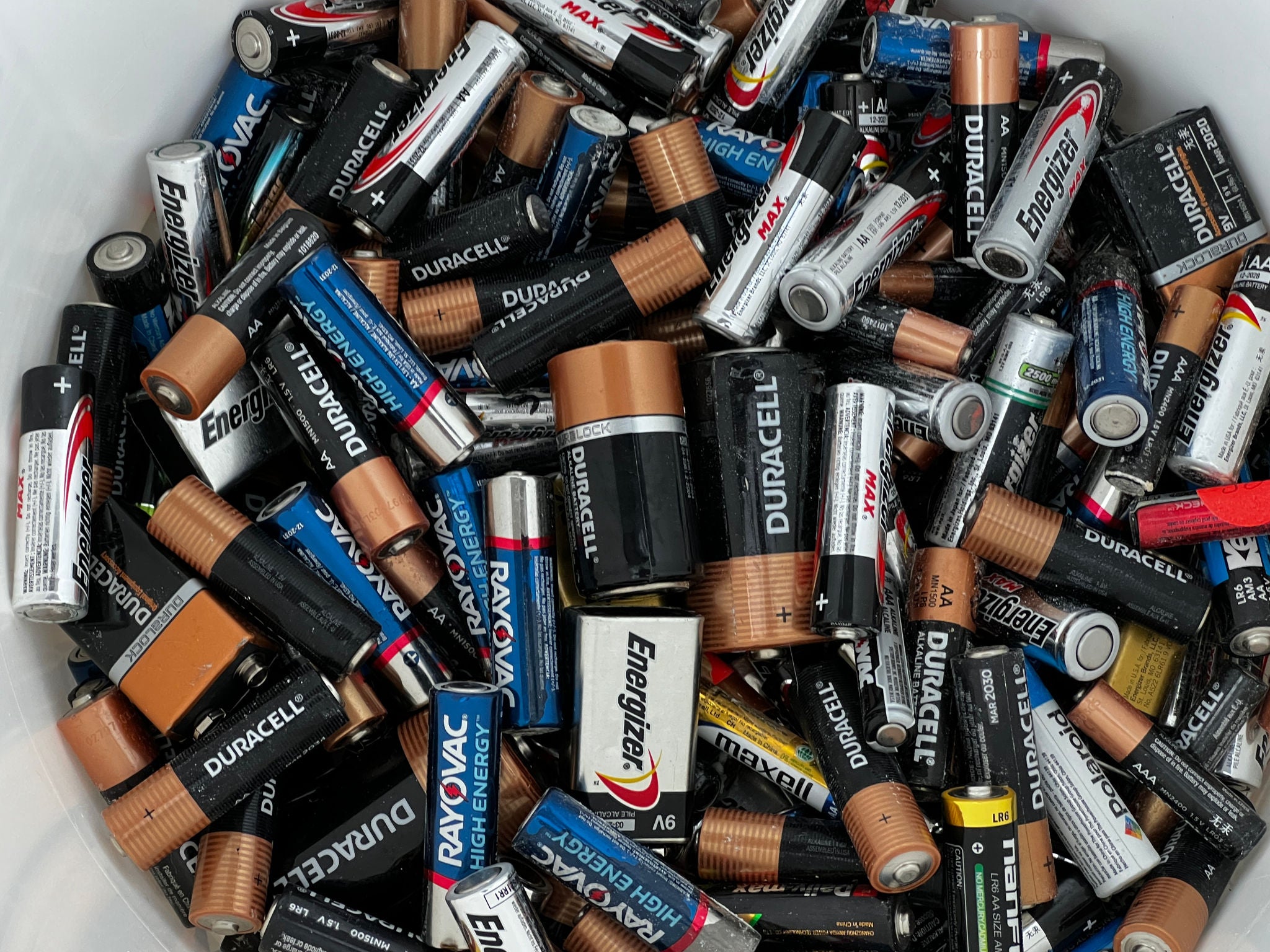 A tight, overhead shot of a white bucket filled with various sizes, shapes, brands, and colors of alkaline batteries.