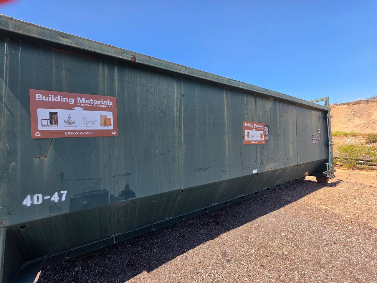 A large, green roll-off dumpster with signs that say "Building Materials." The dumpster is on a gravel surface with a brown hill and blue daylight sky in the background. 
