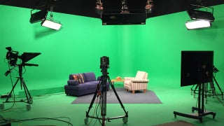 A green screen room set up with cameras, lights, and two seats facing each other.