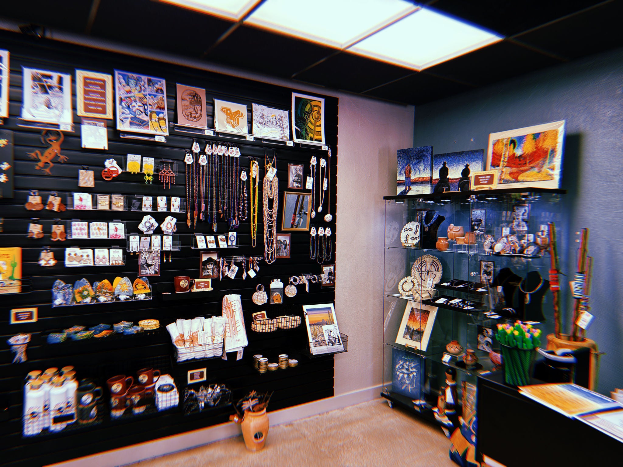 A section of the S’edav Va’aki Museum Store featuring a black slat‑wall display filled with Southwestern and Indigenous‑themed merchandise. The wall is lined with framed art prints at the top, with rows of jewelry, ornaments, magnets, postcards, and small crafts below. Baskets on lower shelves hold folded textiles, jars, and pottery items. To the right, a tall glass display case contains pottery, woven baskets, figurines, jewelry, and framed artwork, with additional art pieces resting on top. The lighting is bright, and the space has neutral flooring and a dark ceiling with fluorescent panels.