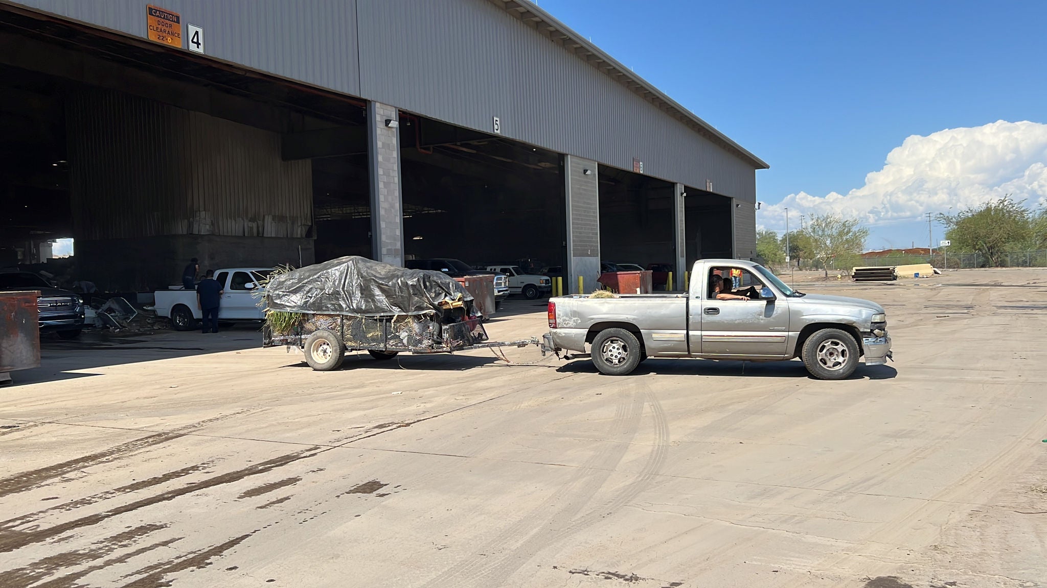 A silver pickup pulling a small trailer with covered garbage backs into a large bay at an industrial facility. There are three other bay doors open in the background, with wide, open space for cars and trucks to maneuver outside. There are some white, puffy clouds against a blue sky on a sunny day.