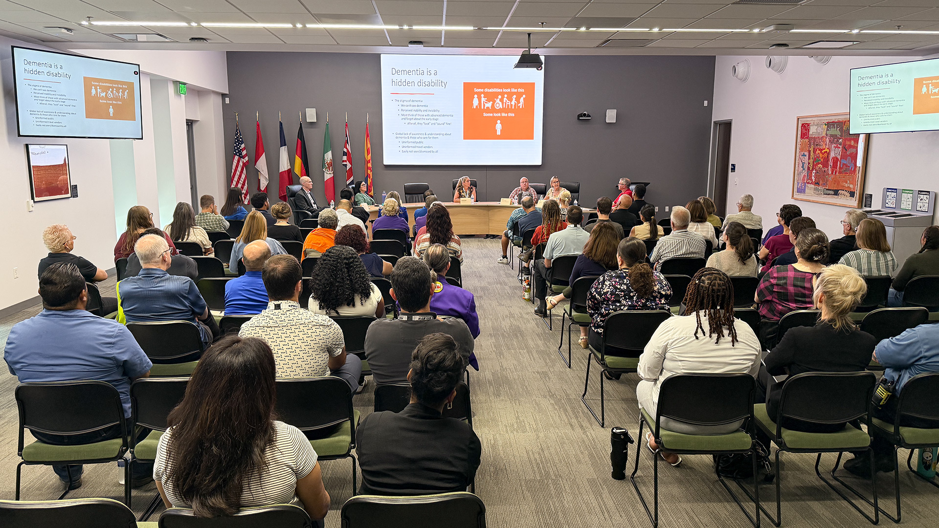 A large group of people attending the Dementia-Friendly Workshop. They are all pacing a panel of speakers and a screen on the wall featuring a presentation.
