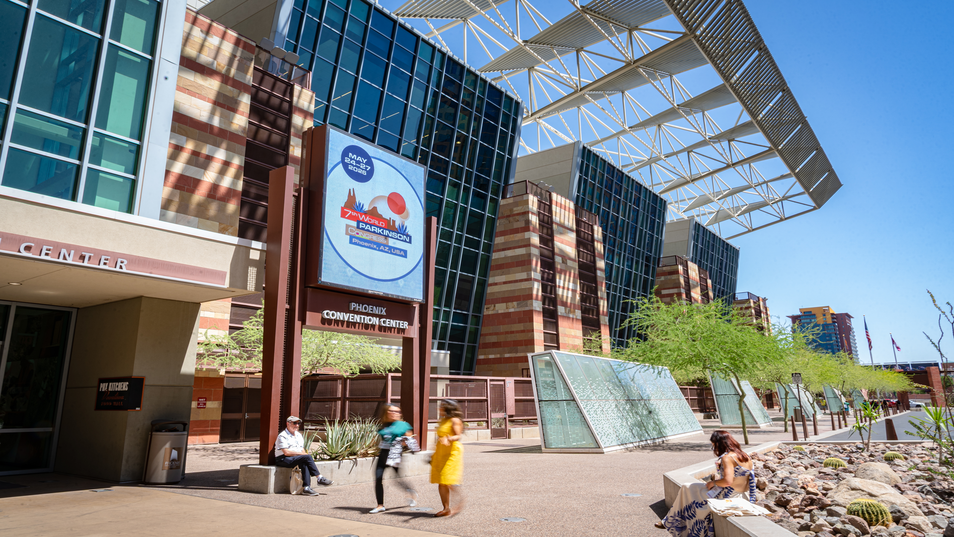 An outside shot of the Phoenix Convention Center showcases an electronic billboard that says 'May 24-27, 2026 | 7th World Parkinson Congress | Phoenix, AZ, USA. A few people are sitting and walking near the entrance to the Convention Center on a sunny day with blue, clear sky. 
