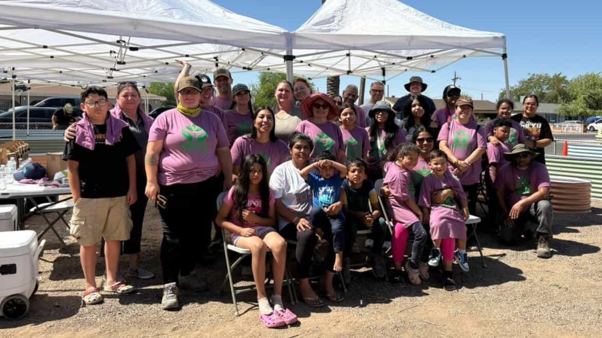 A large group of people, including children and adults, pose for a photo under two white tents. Most are wearing purple neighborhood group t-shirts. The group is outdoors with a dirt ground and some buildings in the background.
