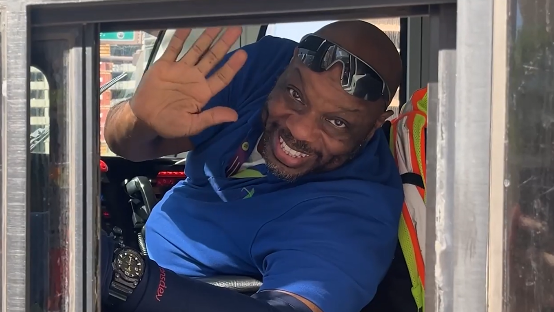 A picture of a light rail operator wearing a blue polo and sunglasses on top of his head, waving and smiling. 