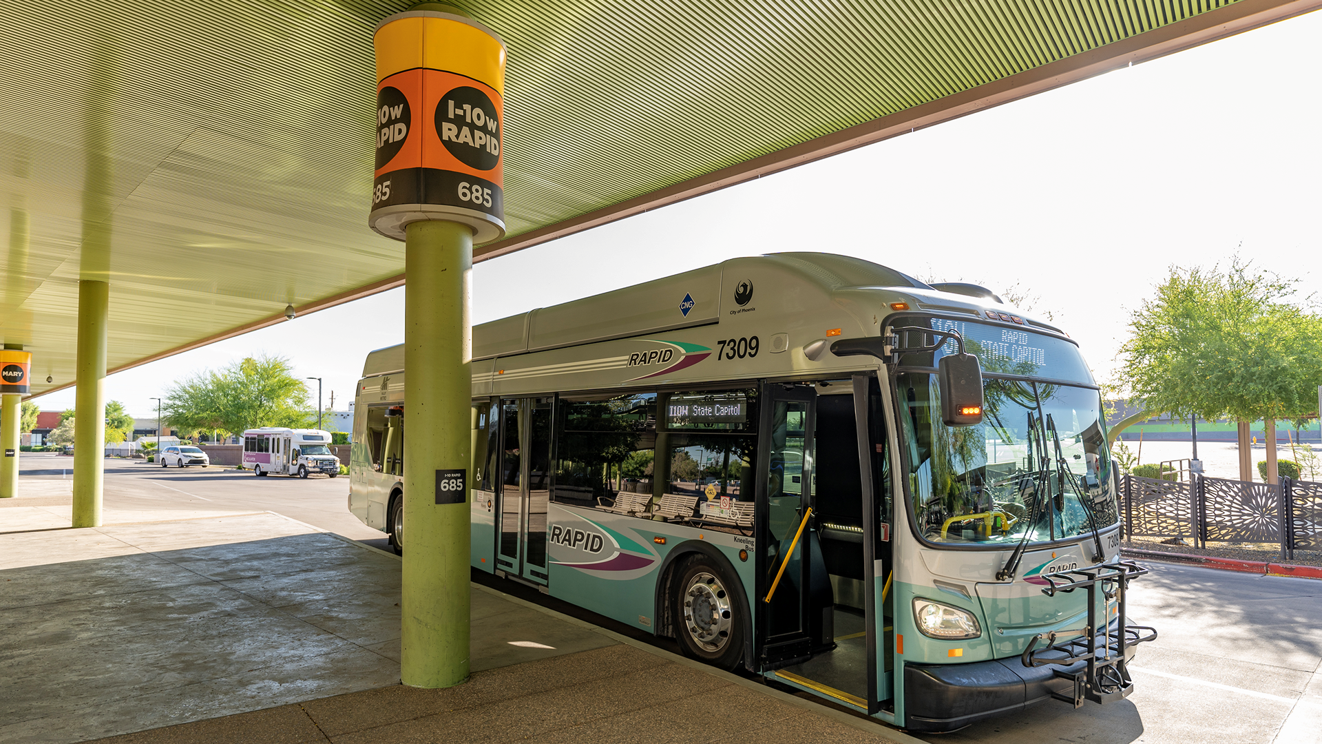 A picture of a silver RAPID bus waiting by its stop at the Desert Sky Transit Center. There are trees and other buses in the background. 
