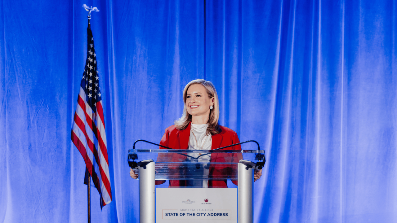 Phoenix Mayor Kate Gallego at lectern with Greater Phoenix Chamber and City of Phoenix logo that says "State of the City Address" in front of American flag and blue curtain backdrop giving State of the City address