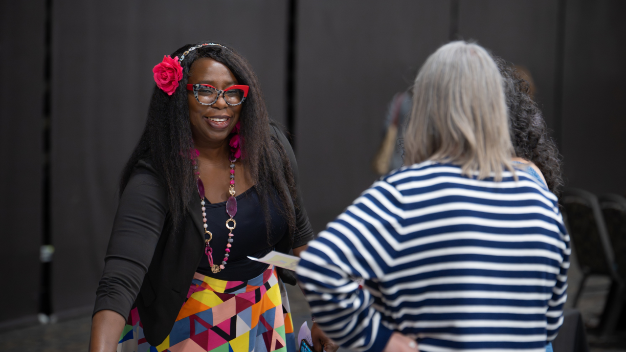 A woman with long dark hair is smiling towards the camera in the background while talking to a woman with a striped long sleeve shirt. The woman smiling is in focus, while the woman in the striped shirt in the foreground has her back to the camera and is out of focus.