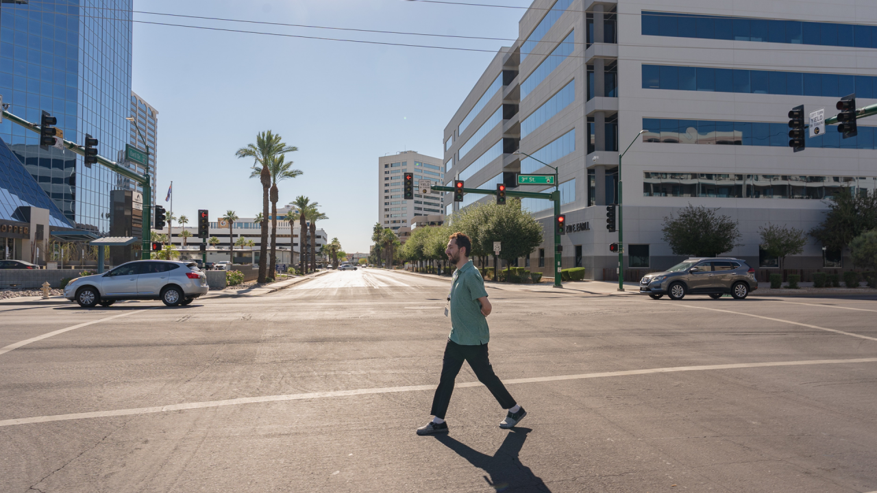 A man in a green shirt walking across a street near 3rd st in Phoenix, with a road and cars in the background. 