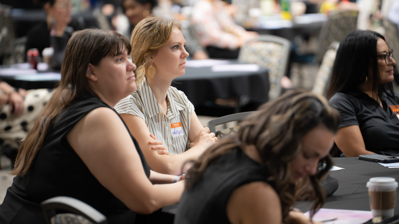 A group of women sitting at a table listening to speaker at a conference. The foreground is out of focus and the background is in focus. 