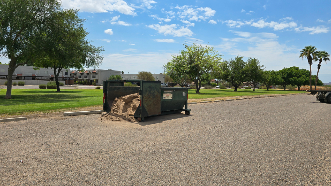 A roll-off metal bin filled with sand its in a parking lot on a sunny day with clouds in a blue sky.