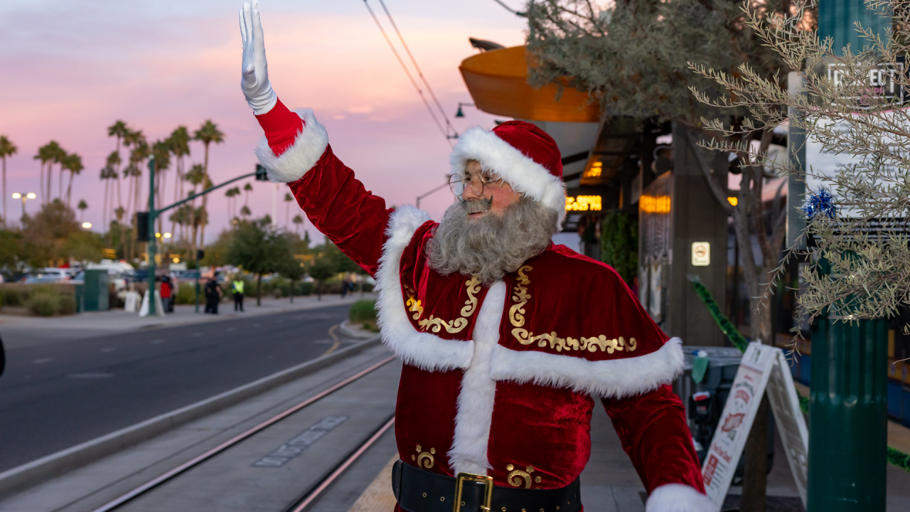 A man dressed as Santa waves at a group out of frame as he waits on the a light rail platform. 