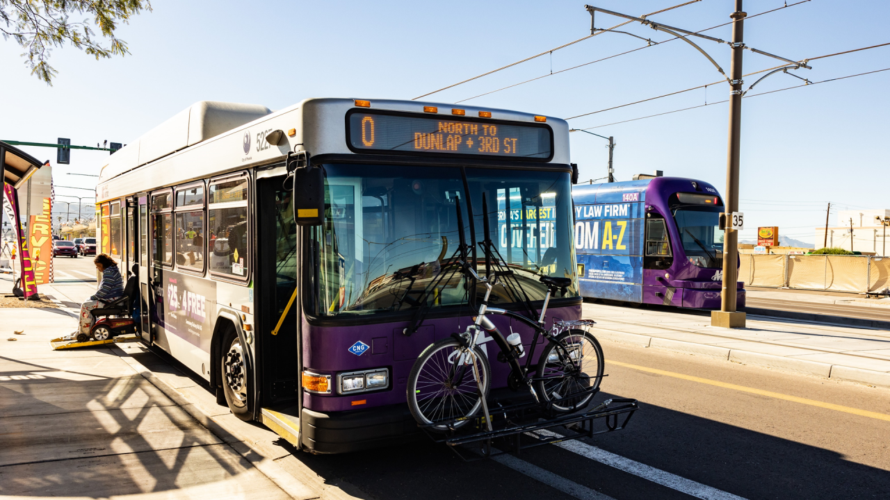 A Valley Metro bus stopped at a bus stop with a person in a wheelchair getting of the bus and a light rail train passing by in the background. 