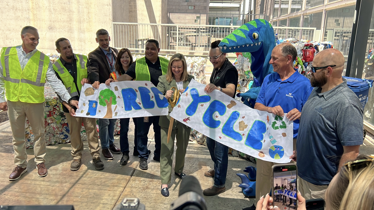 Public Works leaders, and city vendors hold a white, paper banner that features the word 'Recycle' along with children's drawings as Mayor Kate Gallego uses giant scissors for a ceremonial ribbon cutting