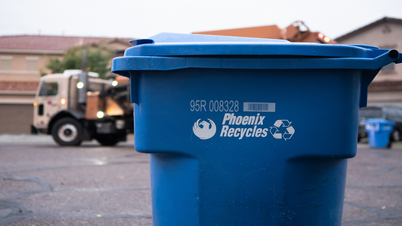 A blue curbside recycle bin that says 'Phoenix Recycles' in white letters with the City of Phoenix bird logo on one side of the text and a recycling logo (three arrows pointing at each other in a circle) on the other. In the background is a garbage truck driving down a residential street on a cloudy day.