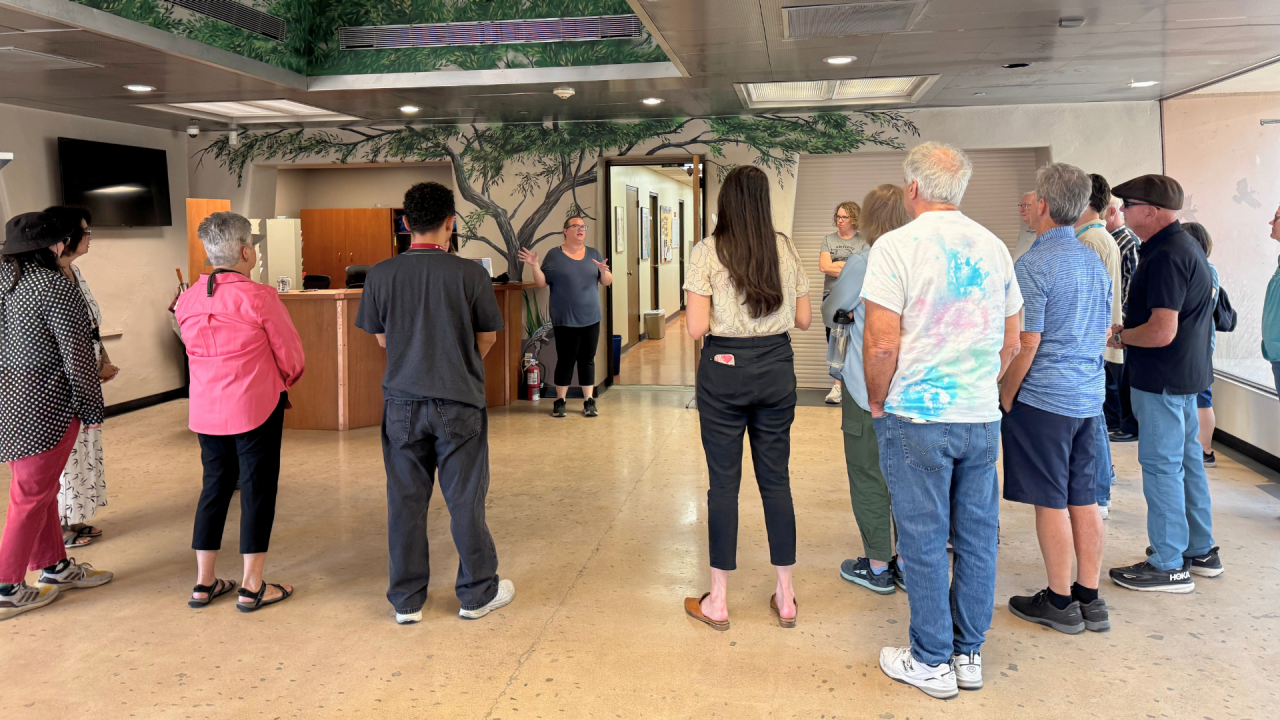 Group photo of volunteers in the S'edav Va'aki Museum Lobby, forming a semi-circle while facing the Curator of Exhibits providing a tour overview. A mural of a mesquite tree is in the backrgound. 